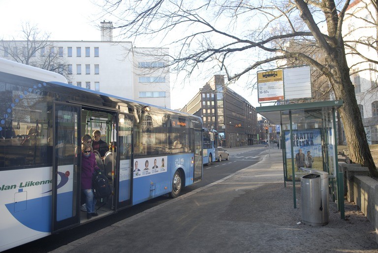 Bussi pysäkillään, taustalla rakennuksia, pysäkin takana puu. Bussista on poistumassa ihminen aurinkolaseissa, punaisessa takissa ja farkuissa.