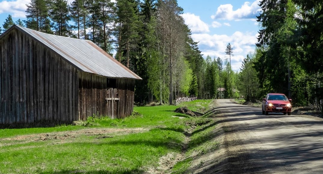 Kuvituskuva artikkeliin, jonka aiheena on maaseudun elinvoima, ilmastonmuutos ja ruokaturva. Punainen auto ajaa metsäisessä maisemassa harmaan ladon ohitse tiellä maaseudulla.