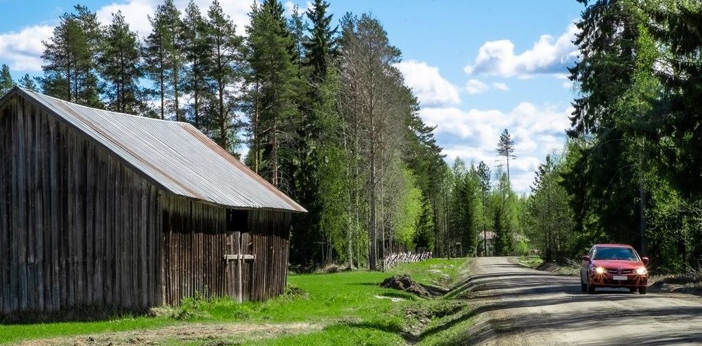 Kuvituskuva artikkeliin, jonka aiheena on maaseudun elinvoima, ilmastonmuutos ja ruokaturva. Punainen auto ajaa metsäisessä maisemassa harmaan ladon ohitse tiellä maaseudulla.